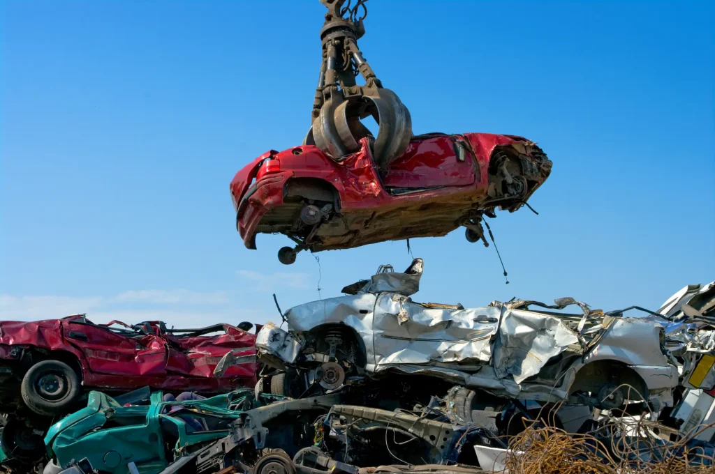 A crane grab lifting a crushed red car above a pile of scrap vehicles