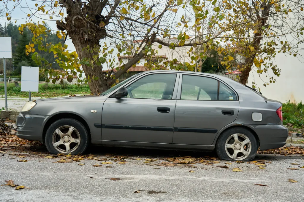 An old grey saloon with flat tyres parked under autumn trees on a leaf-covered street