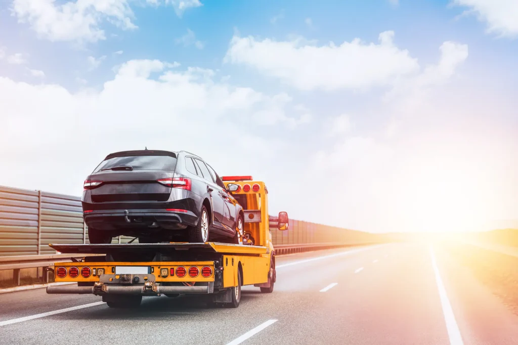 A recovery truck carrying a dark hatchback along a motorway in bright sunshine